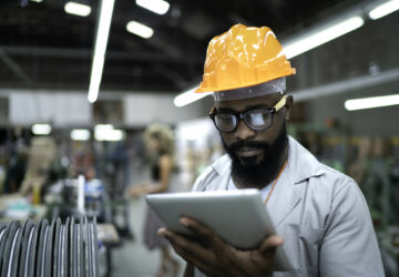 A man wearing a hardhat reviews information on a tablet while in a factory.