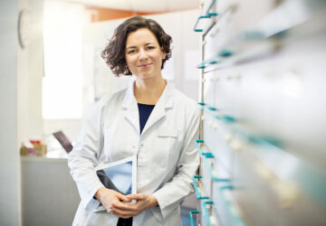 Portrait of confident young female pharmacist leaning to a medicine shelf with a digital tablet.